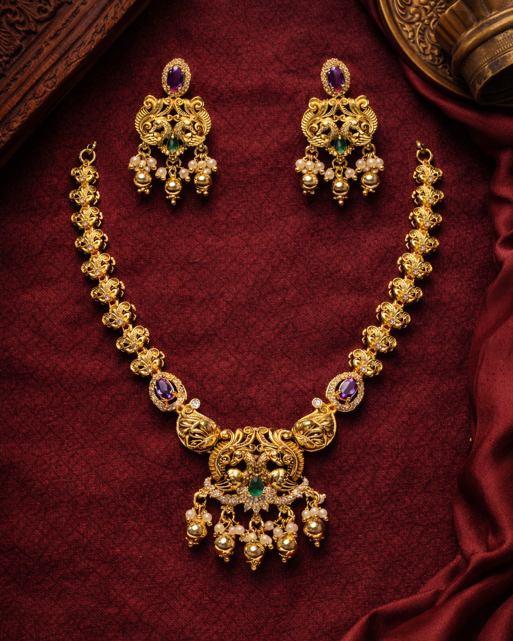Gold necklace with green and purple stones on a red fabric background suwarnikaa jewels www.suwarnikaajewels.com 