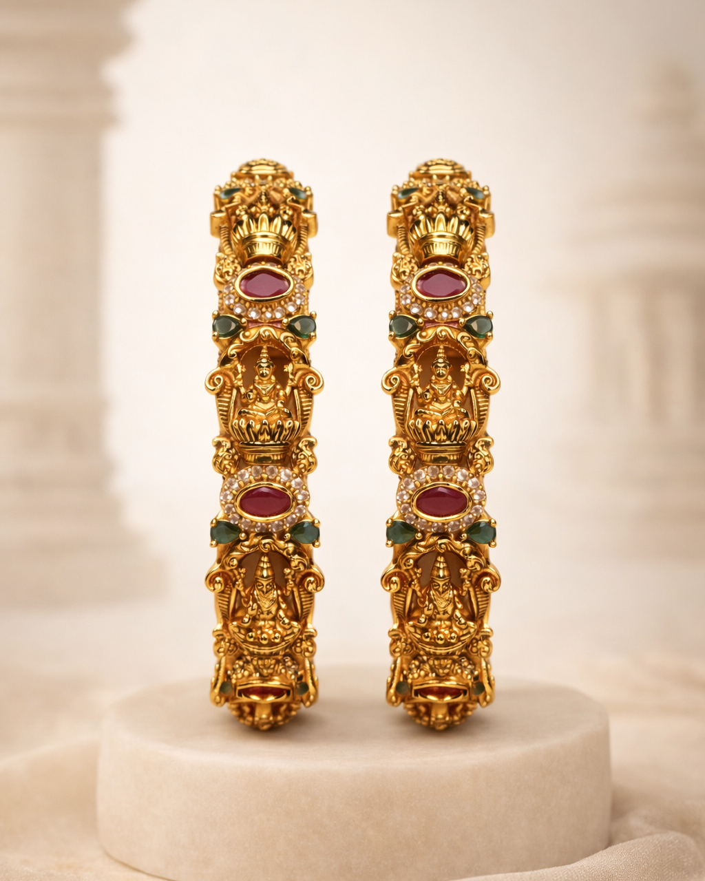 Pair of gold earrings with red and green gemstones on a beige SRI LAKSHMI VALAYAAM BY SUWARNIKAA JEWELSbackground